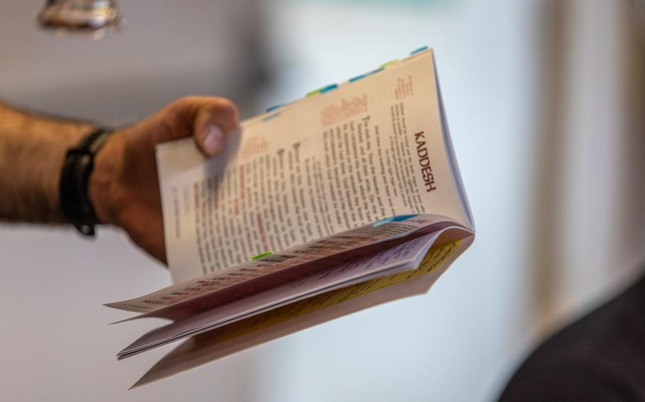 A close-up of a person’s hand holding a book with page markers, opened to a page titled “KADESH.” The background is softly blurred, focusing attention on the booklet.