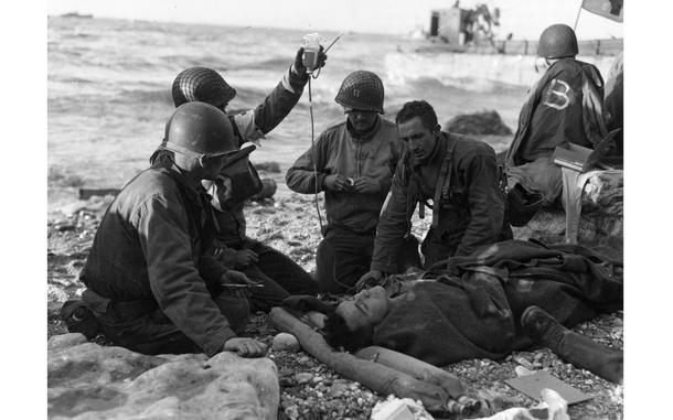 Nurses administer a plasma transfusion to a wounded survivor of a landing craft at "Fox Green" sector portion of Omaha Beach. The photograph shows how promptly medical treatment was administered on the beach. Note the inflatable life belt used as a pillow, and the feet of second prostrate man on (right). 5th Engineer Special Brigade. The flag on the far right is a flag of the Red Cross.   The Soldiers are from the 16th Infantry Regiment ,1st Infantry Division. The exact date of the image is unknow as sources differ on it and both June 6th (D-Day) as well as June 12th is given.