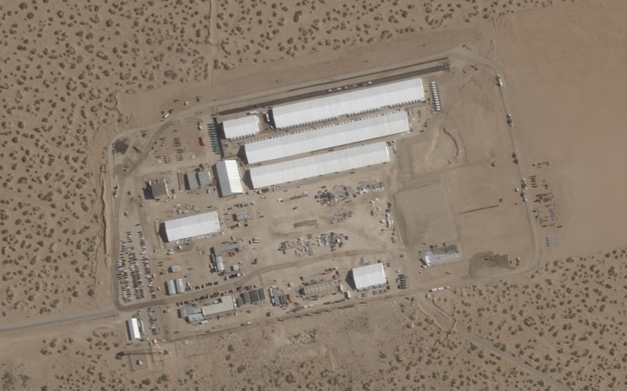 An overhead satellite image shows long, rectangular white and gray buildings in a desert setting.
