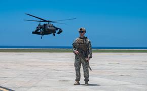 A Marine in camouflage combat gear stands on a tarmac with a helicopter hovering above the ground behind him and the horizon in the background.