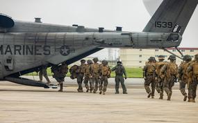 A line of Marines in camouflage combat gear walk toward the back of an aircraft on a runway and up a ramp to board.