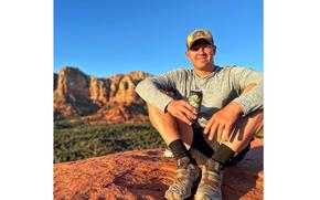 Reed sits on a rock with a drink can in his hand, mountains and blue skies in the background.