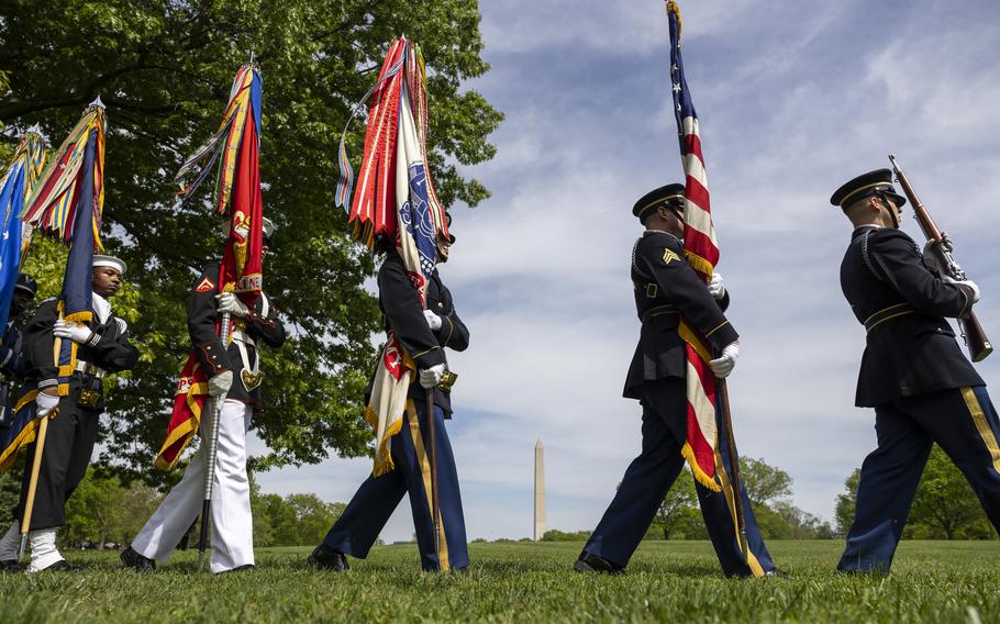 A service member holding a rifle leads other service members holding flags as they march single file through a patch of grass.