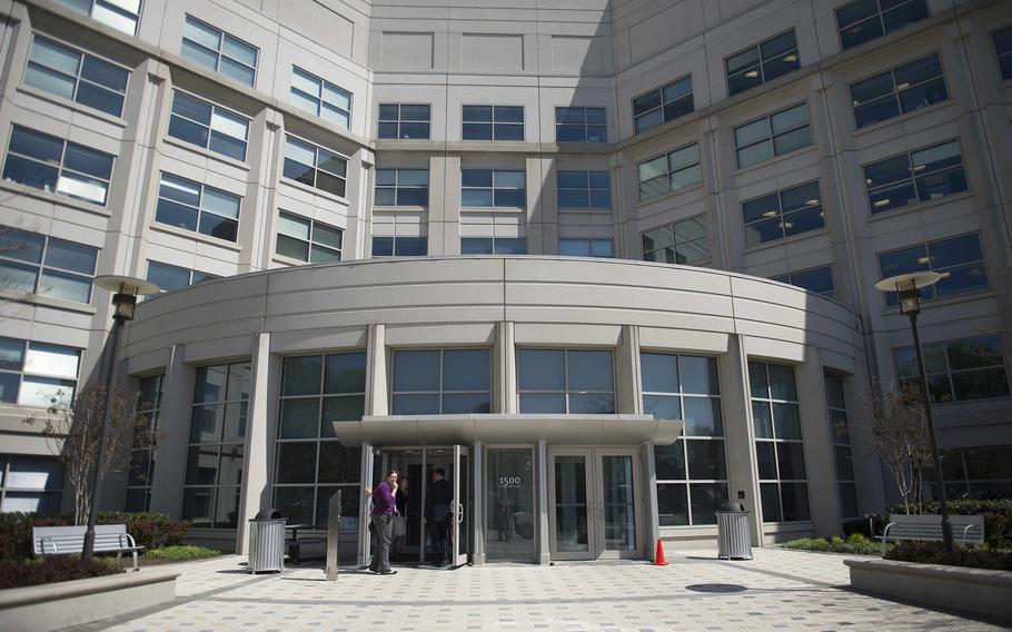 The front entrance of a multi-story government office building is seen, with a man standing in front of one of three sets of glass double doors.