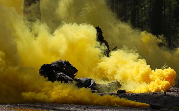 Two soldiers in tactical gear move through a dense cloud of yellow smoke in a wooded area. One soldier is prone on the ground in the foreground, while another is partially visible in the background. Multiple smoke canisters are emitting thick yellow smoke, obscuring much of the scene. 
