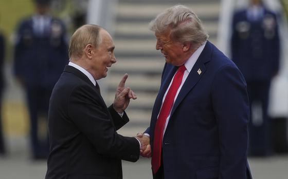 Donald Trump and Vladimir Putin, both wearing dark suits, shake hands on an airfield runway with the blurred stairs of an airplane in the background.