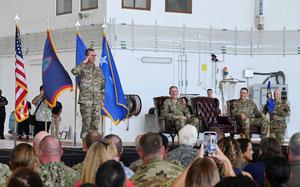 A uniformed officer stands at attention and salutes in front of four flags, including the U.S. flag and the Guam territorial flag. Three other military officers, also in uniform, are seated on a stage with leather chairs, smiling and observing the proceedings. 