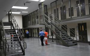 A guard in blue uniform escorts a detainee in orange jumpsuit past a set of steel stairs through a detention facility.