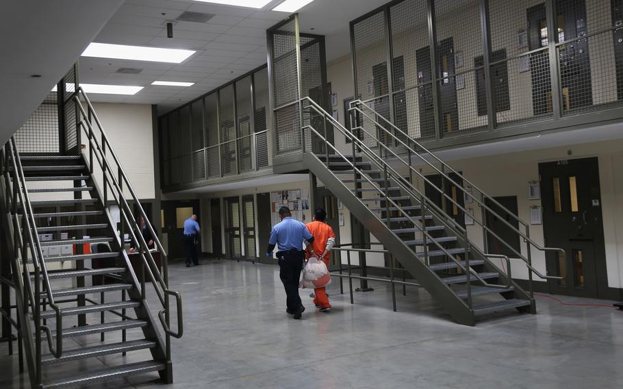 A guard in blue uniform escorts a detainee in orange jumpsuit past a set of steel stairs through a detention facility.