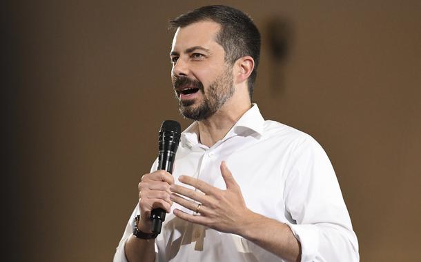 Pete Buttigieg, wearing a white button-down shirt, holds a microphone and speaks on a stage.