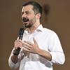 Pete Buttigieg, wearing a white button-down shirt, holds a microphone and speaks on a stage.