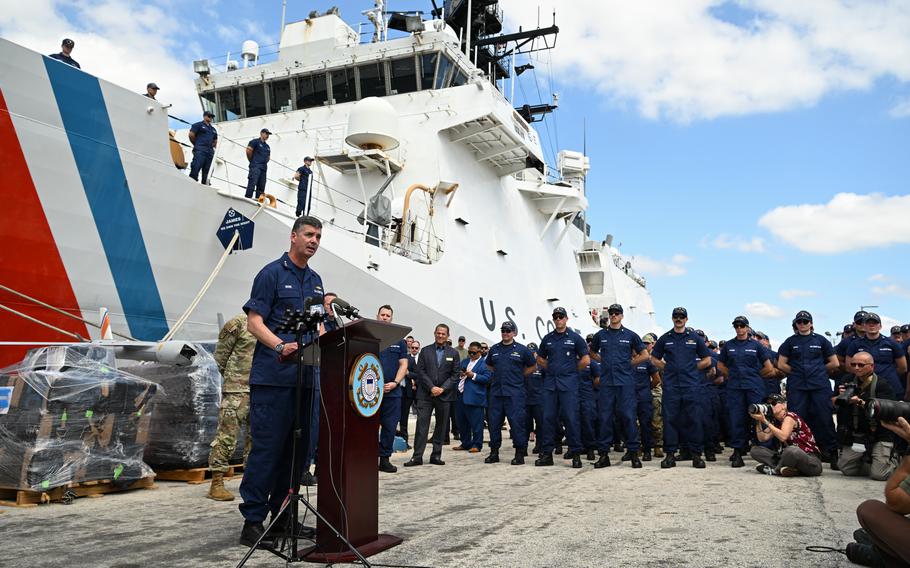 Moore speaks at a microphone with a Coast Guard crew and cutter in the background.