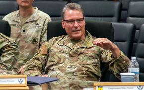 A military officer in camouflage uniform and dark-rimmed glasses gestures with his left hand while seated at a conference table.