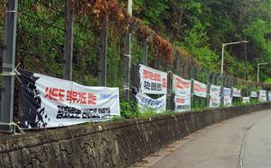 White banners with red and black Korean-language writing hang on a chain link fence atop a small stone wall, with dense green vegetation behind it.