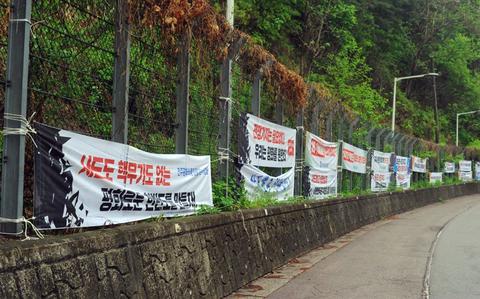White banners with red and black Korean-language writing hang on a chain link fence atop a small stone wall, with dense green vegetation behind it.