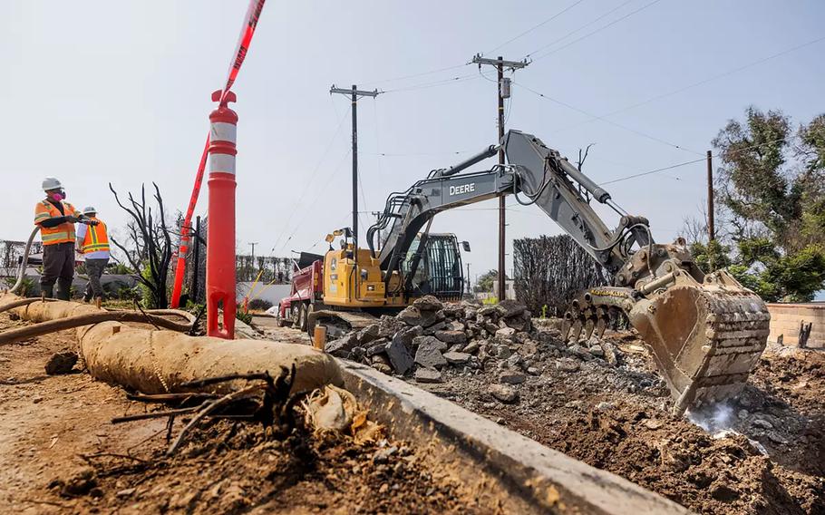 Contractors operate a backhoe to clear debris from a property damaged by wildfire.