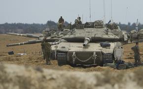 Soldiers in camouflage sit on and stand next to tanks in a sandy desert setting.