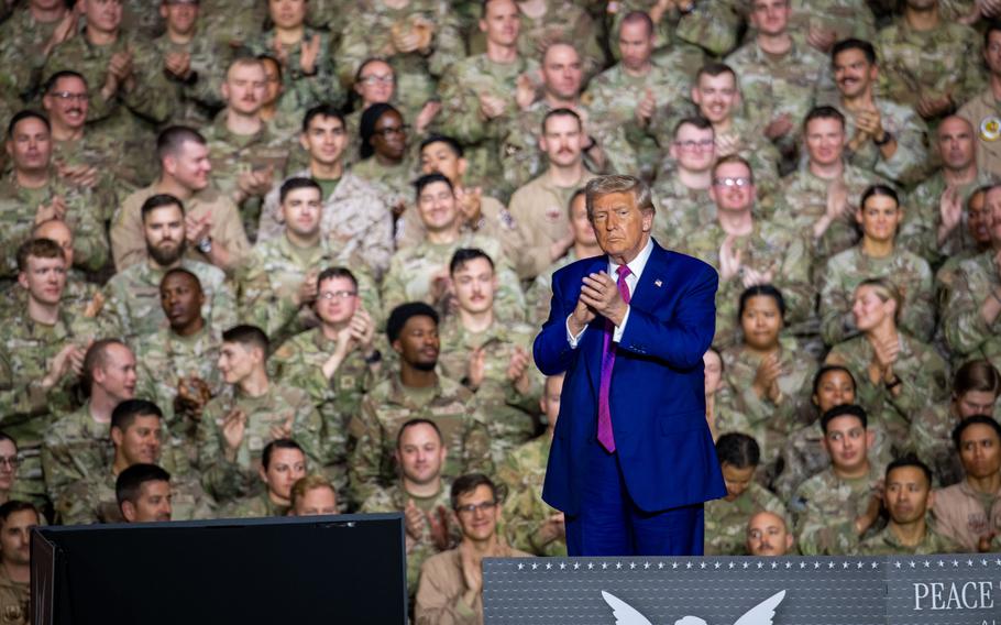 President Donald Trump in a blue suit, clapping his hands on a stage while soldiers watch behind him.