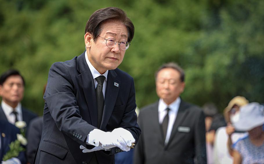 A man in a black suit and white gloves, wearing glasses and a South Korean flag pin, participates in a solemn memorial ceremony outdoors. He is surrounded by other mourners dressed in formal black attire, some holding white flowers. 