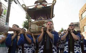 A group of men in traditional blue and white patterned Japanese festival clothing (happi coats) carry a portable shrine (mikoshi) on their shoulders during a street festival. The scene takes place on a city street with buildings and power lines visible in the background.