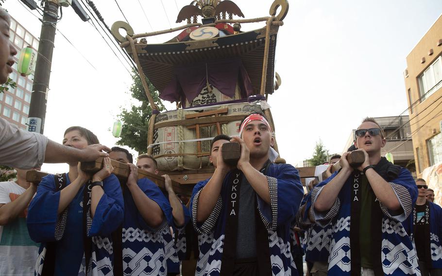 A group of men in traditional blue and white patterned Japanese festival clothing (happi coats) carry a portable shrine (mikoshi) on their shoulders during a street festival. The scene takes place on a city street with buildings and power lines visible in the background.