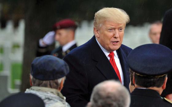 President Donald Trump speaks to a group of people gathered in front of him. A service member standing in front of cemetery crosses salutes in the background. 