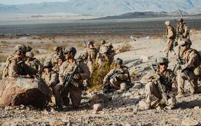 A group of military personnel in desert camouflage uniforms and tactical gear are positioned on rocky, arid terrain with rifles and equipment. 