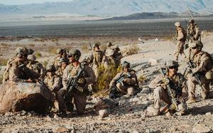 A group of military personnel in desert camouflage uniforms and tactical gear are positioned on rocky, arid terrain with rifles and equipment. 