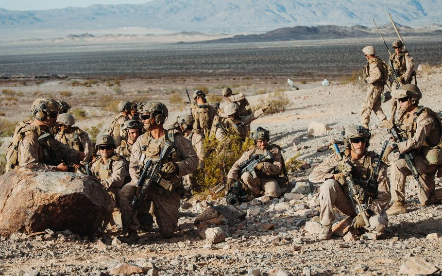 A group of military personnel in desert camouflage uniforms and tactical gear are positioned on rocky, arid terrain with rifles and equipment. 