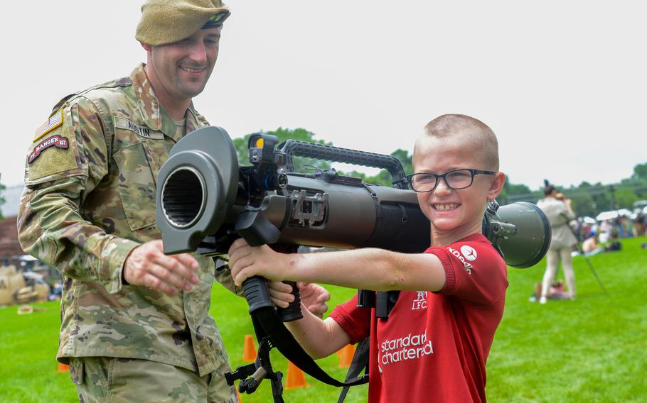 A young boy smiles as he holds a weapon and a soldier watches.
