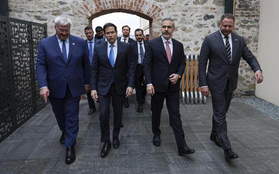 Four government officials in suits walk through a corridor with a stone wall and entrance way behind them.