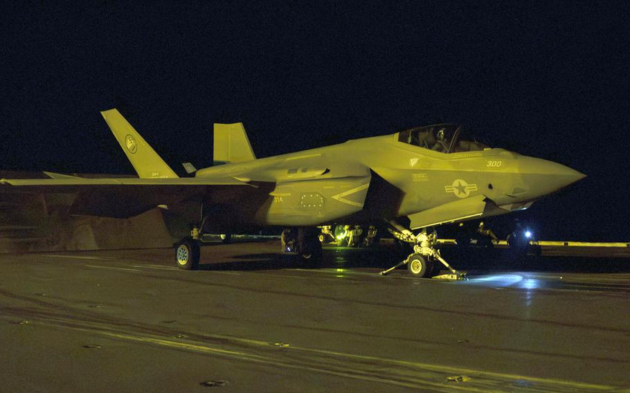 A military fighter jet sits on the deck of an aircraft carrier at night as it prepares to take off.