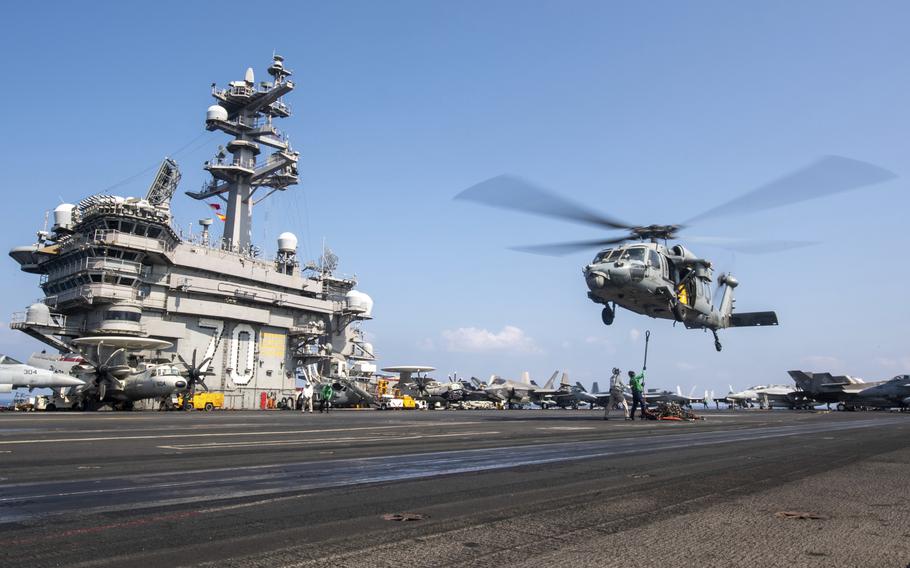 A military helicopter lands on the deck of an aircraft carrier, with the bridge of the ship visible to the left.