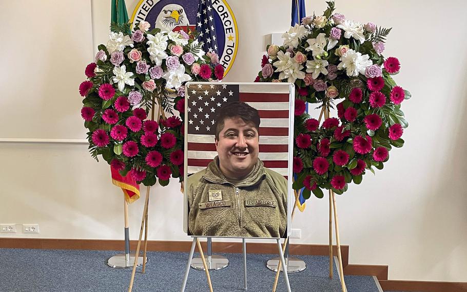 A large photo of a U.S. airman sits on an easel flanked by two wreaths of red and white flowers during a memorial service.
