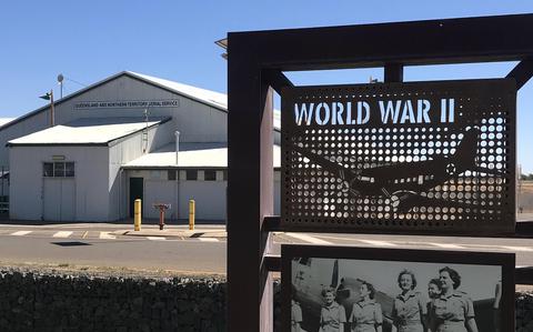 A World War II memorial display featuring a perforated metal panel with “WORLD WAR II” text and a silhouette of an aircraft, positioned in front of a large hangar building labeled “Queensland and Northern Territory Aerial Service.” A historical photograph of four women in military uniforms is mounted below the main panel.