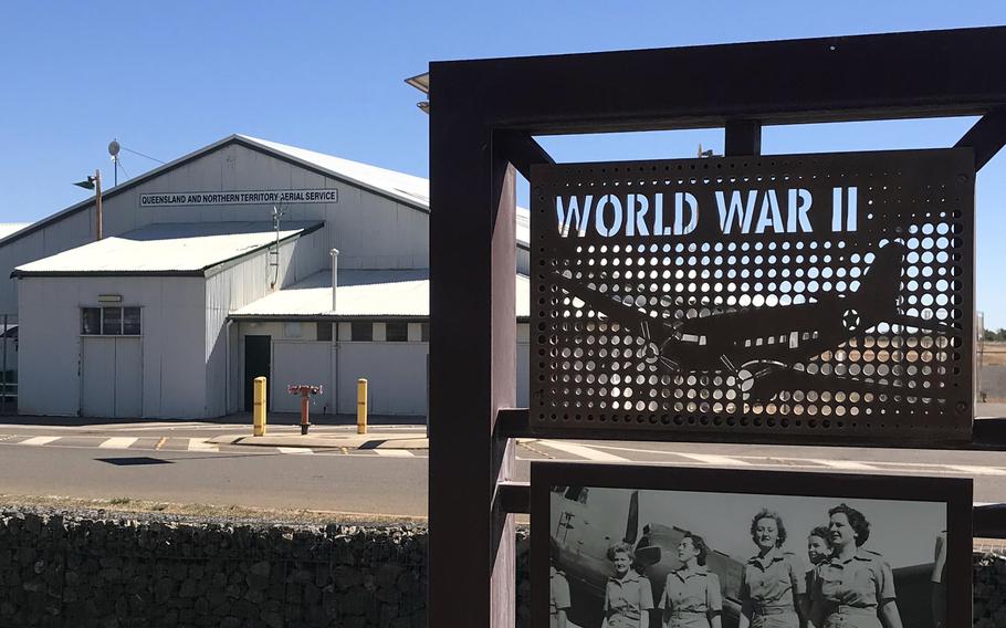 A World War II memorial display featuring a perforated metal panel with “WORLD WAR II” text and a silhouette of an aircraft, positioned in front of a large hangar building labeled “Queensland and Northern Territory Aerial Service.” A historical photograph of four women in military uniforms is mounted below the main panel.