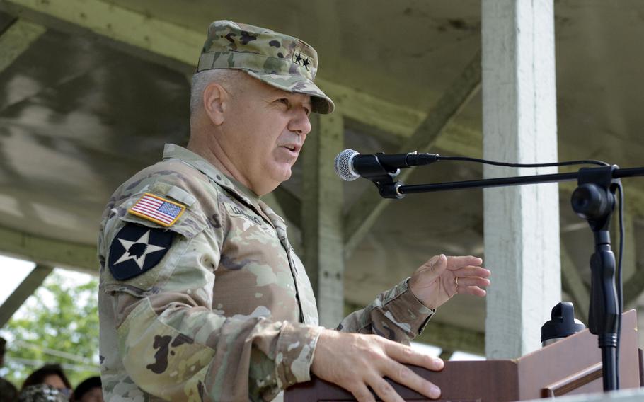 An Army officer in camouflage uniform and hat stands at a podium and speaks into a microphone under a wooden pavilion.