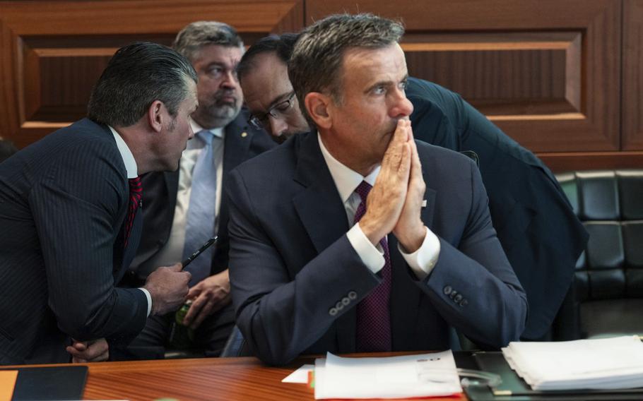A man in a suit claspses his hands together while eated at a table with blurred white pages in front of him.