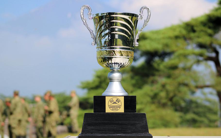 A shiny silver and gold trophy labeled “Garrison Games” stands on a wooden platform outdoors, with blurred service members in camouflage and green trees in the background.