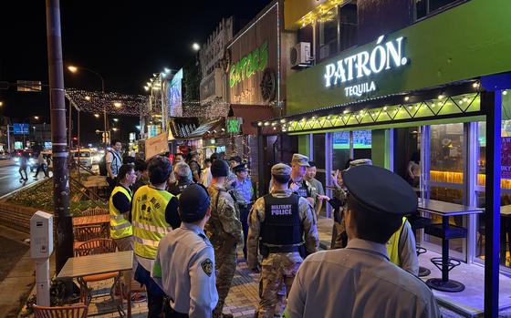 A group of military police, local law enforcement and security personnel are gathered outside a brightly lit bar named “PATRÓN Tequila” on a busy street at night. The officers appear to be conducting a joint patrol with several wearing reflective vests and uniforms. Traffic and pedestrians are visible in the background under streetlights.