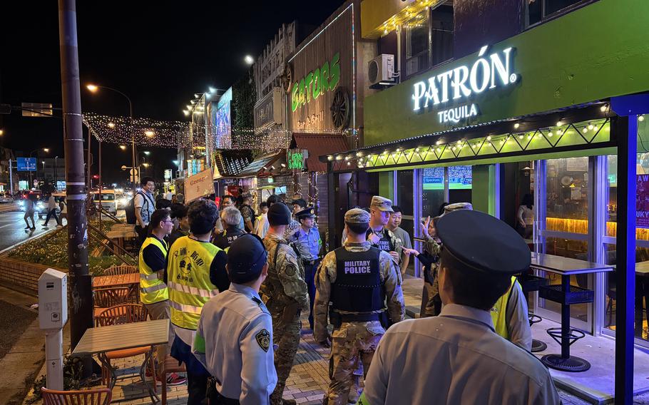 A group of military police, local law enforcement and security personnel are gathered outside a brightly lit bar named “PATRÓN Tequila” on a busy street at night. The officers appear to be conducting a joint patrol with several wearing reflective vests and uniforms. Traffic and pedestrians are visible in the background under streetlights.