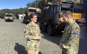 A U.S. Army soldier in camouflage uniform with glasses and curly hair shakes hands with an Australian military officer wearing the Australian flag patch on his uniform. They are standing on a cobblestone surface in front of large military vehicles.
