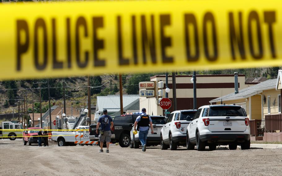 Police crime scene with yellow police tape in the foreground stretches across the frame. Behind the tape, two people in police vests walk among several white police SUVs parked on a dirt road in what appears to be a small rural town.