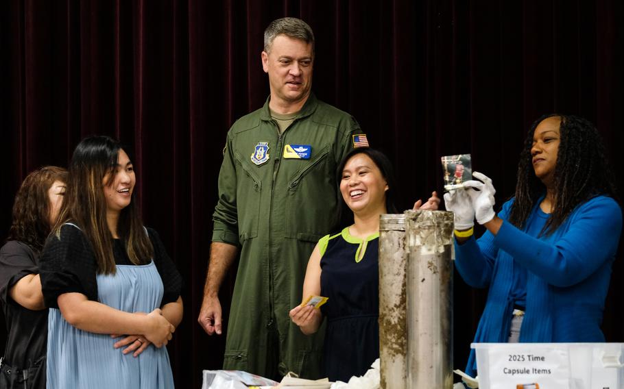 A woman in gloves holds up a photograph as adults open a silver time capsule that’s sitting on a table. 