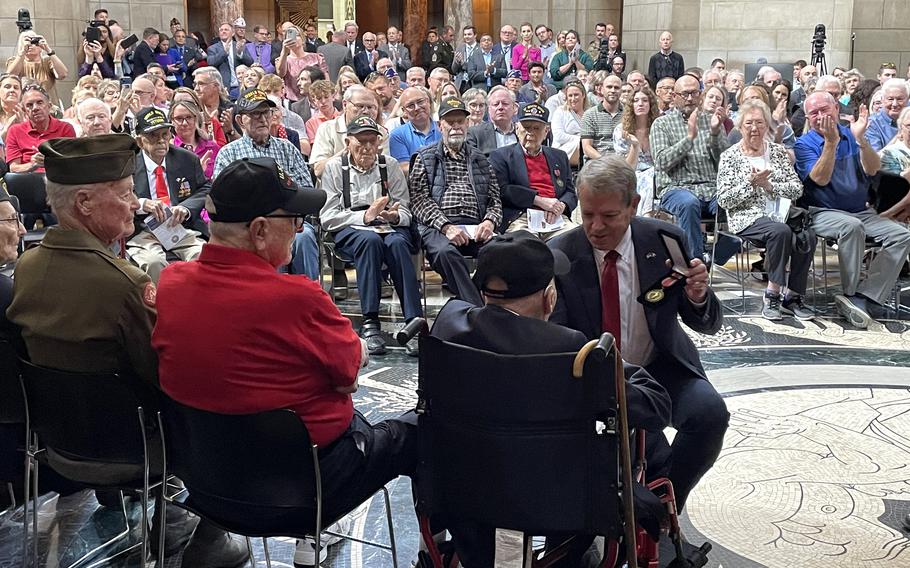 A man in a suit crouches with a medal in one hand, near elderly veterans seated in front of him.