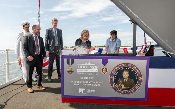 Several people, some in suits and one in a white Navy uniform, smiling as a champagne bottle is opened or smashed.