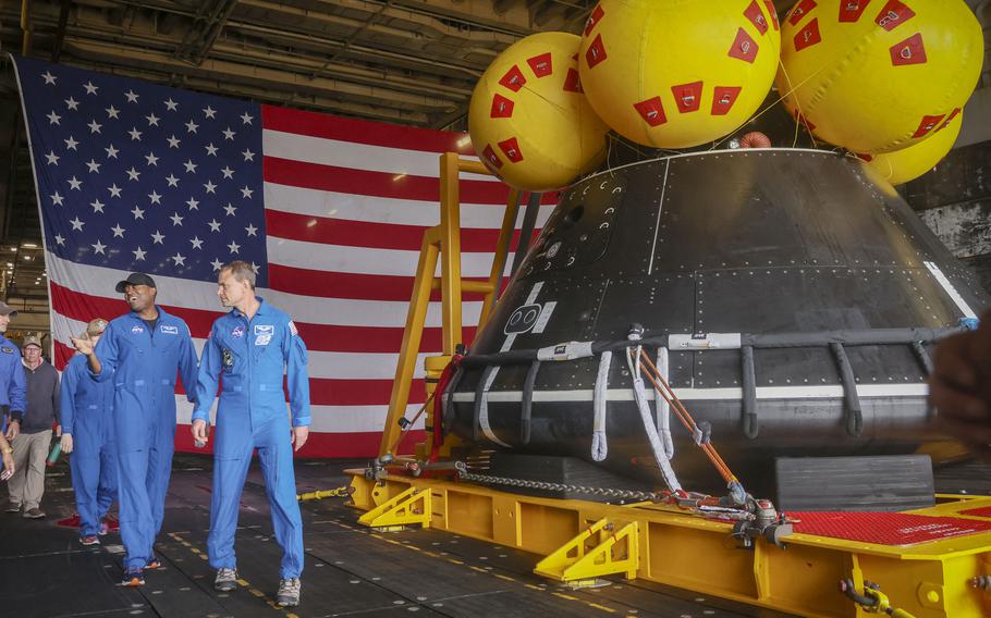 Astronauts walk next to a spacecraft with a large American flag in the background.