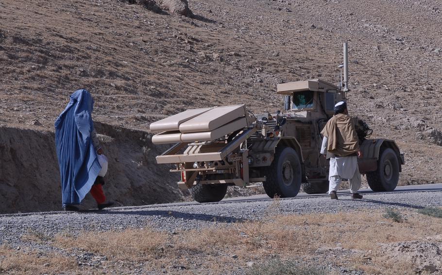 Two civilians walk near a military vehicle in a hilly area.