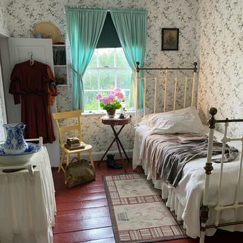 A replica setting of an early 20th-century bedroom. A single bed is placed against the wall on the right, with a small side table next to it with a case of pink flowers and a red dress hanging from a closet door on the left.