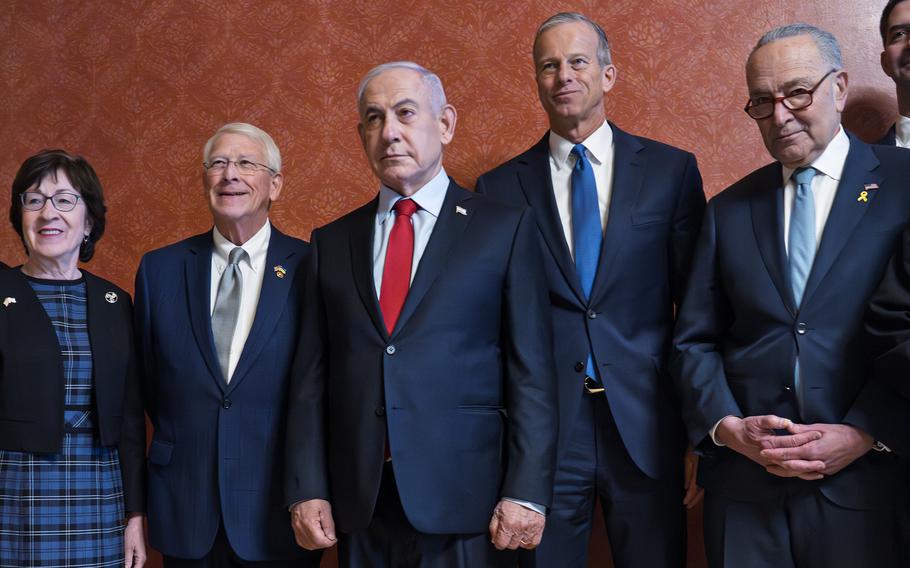 Benjamin Netanyahu, wearing a dark suit and red tie, poses with a group of senators in front of an orange-patterned wall in the U.S. Capitol building.